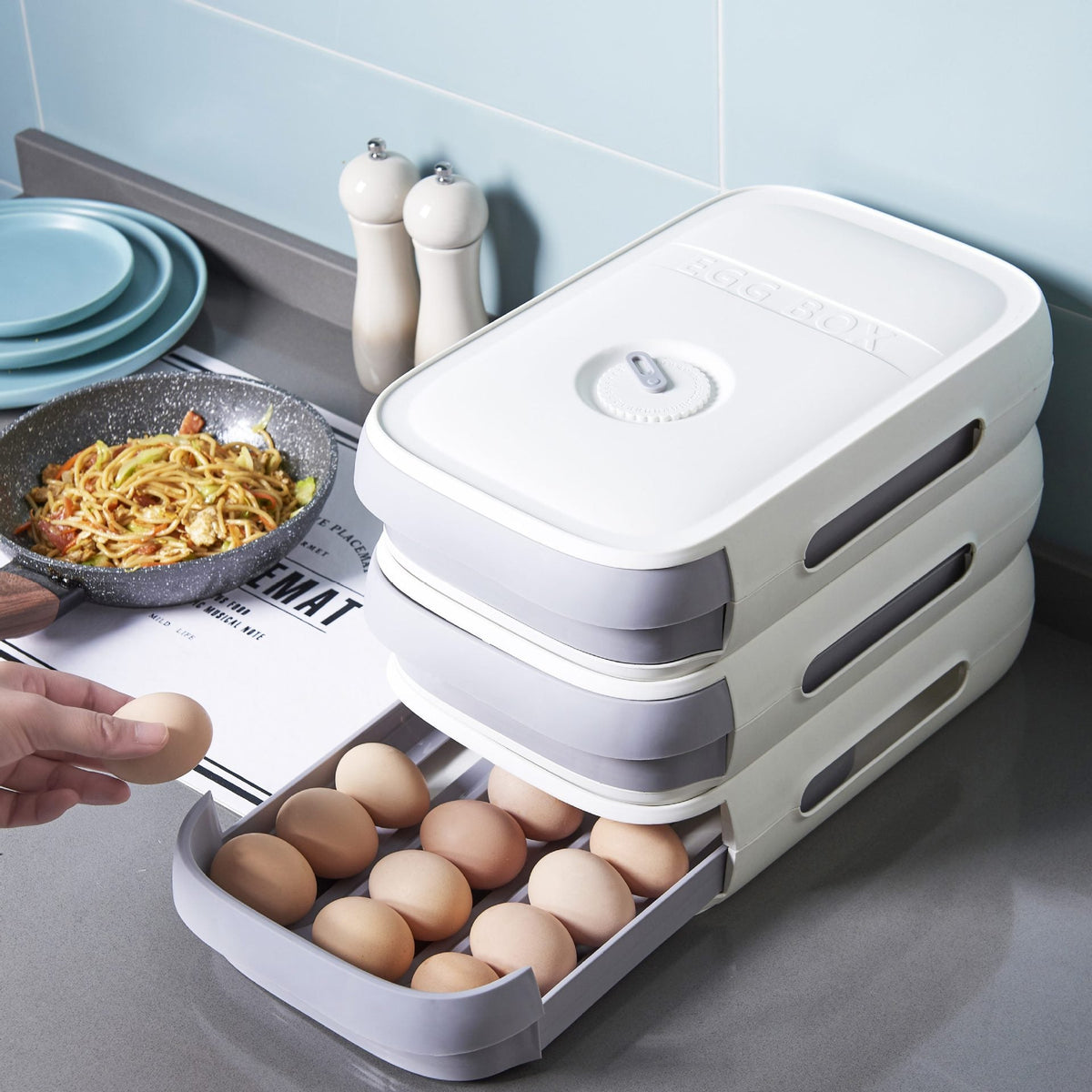 Three white and gray egg organizer units stacked on a kitchen counter, with the bottom drawer open showing eggs.