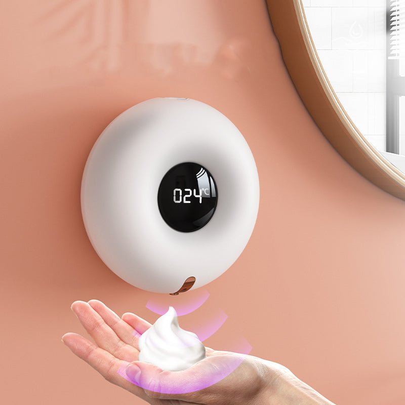 Round white Foamora wall-mounted soap dispenser with a black display, shown dispensing foam into a hand beside a mirror.